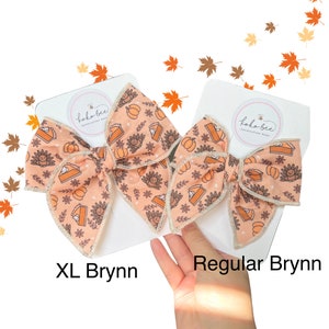 May include: Two hair bows with a Thanksgiving theme. The bows are made of a peach-colored fabric with a pattern of pumpkins, pies, and turkeys. The larger bow is labeled "XL Bryn", and the smaller bow is labeled "Regular Bryn".