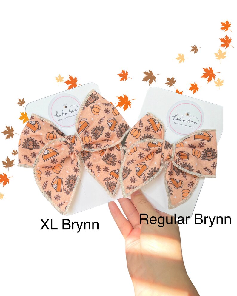 May include: Two hair bows with a Thanksgiving theme. The bows are made of a peach-colored fabric with a pattern of pumpkins, pies, and turkeys. The larger bow is labeled "XL Bryn", and the smaller bow is labeled "Regular Bryn".