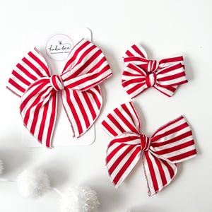 May include: Three red and white striped fabric hair bows of different sizes. The largest bow is on a white card with the text "Koko Bee - Handcrafted Hair Bows".