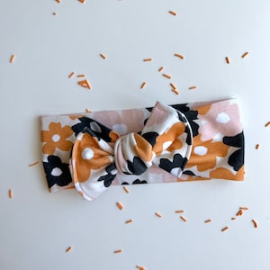 Op de afbeelding: Een babyhoofdband met bloemenpatroon en een strik. De hoofdband heeft een herhalend patroon van zwarte, witte en mosterdkleurige bloemen op een lichtroze achtergrond. De strik is in het midden geplaatst.