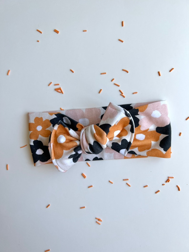 Op de afbeelding: Een babyhoofdband met bloemenpatroon en een strik. De hoofdband heeft een herhalend patroon van zwarte, witte en mosterdkleurige bloemen op een lichtroze achtergrond. De strik is in het midden geplaatst.