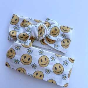 Pode incluir: Duas faixas de cabelo brancas com um padr&atilde;o repetido de rostos sorridentes amarelos e pequenas margaridas brancas. Uma faixa &eacute; amarrada em um la&ccedil;o, e a outra &eacute; uma faixa simples. As faixas de cabelo s&atilde;o feitas de tecido macio.