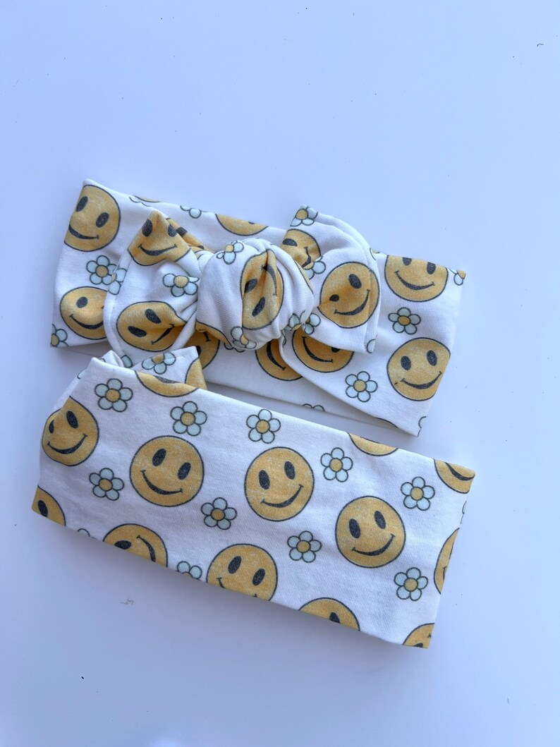 Pode incluir: Duas faixas de cabelo brancas com um padr&atilde;o repetido de rostos sorridentes amarelos e pequenas margaridas brancas. Uma faixa &eacute; amarrada em um la&ccedil;o, e a outra &eacute; uma faixa simples. As faixas de cabelo s&atilde;o feitas de tecido macio.