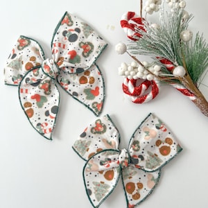 May include: Two green and white Christmas-themed hair bows with a gingerbread man, candy cane, and Christmas tree pattern. The bows are tied with a knot in the center.