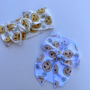 Pode incluir: Duas faixas de cabelo brancas com um padr&atilde;o repetido de rostos sorridentes amarelos e pequenas flores de margarida. Uma &eacute; amarrada em um la&ccedil;o. A outra &eacute; uma faixa larga. As faixas de cabelo est&atilde;o em uma superf&iacute;cie branca.