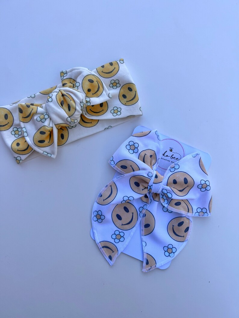 Pode incluir: Duas faixas de cabelo brancas com um padr&atilde;o repetido de rostos sorridentes amarelos e pequenas flores de margarida. Uma &eacute; amarrada em um la&ccedil;o. A outra &eacute; uma faixa larga. As faixas de cabelo est&atilde;o em uma superf&iacute;cie branca.