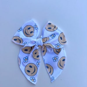 Pode incluir: La&ccedil;o de tecido branco com um padr&atilde;o repetido de rostos sorridentes e pequenas flores de margarida em bege e azul marinho. O la&ccedil;o &eacute; amarrado no centro e tem pontas longas e pontiagudas.