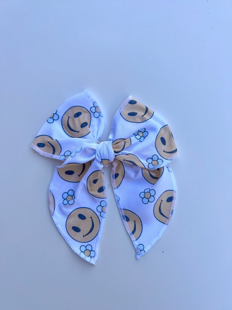 Pode incluir: La&ccedil;o de tecido branco com um padr&atilde;o repetido de rostos sorridentes e pequenas flores de margarida em bege e azul marinho. O la&ccedil;o &eacute; amarrado no centro e tem pontas longas e pontiagudas.