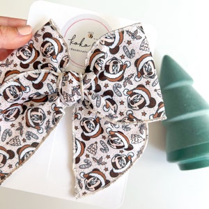 May include: A white fabric bow with a Christmas-themed Minnie Mouse print. The bow is tied with a white ribbon and has a stitched edge. The bow is on a white background with a small circle that says "Koko Handcraft" with a bee logo.