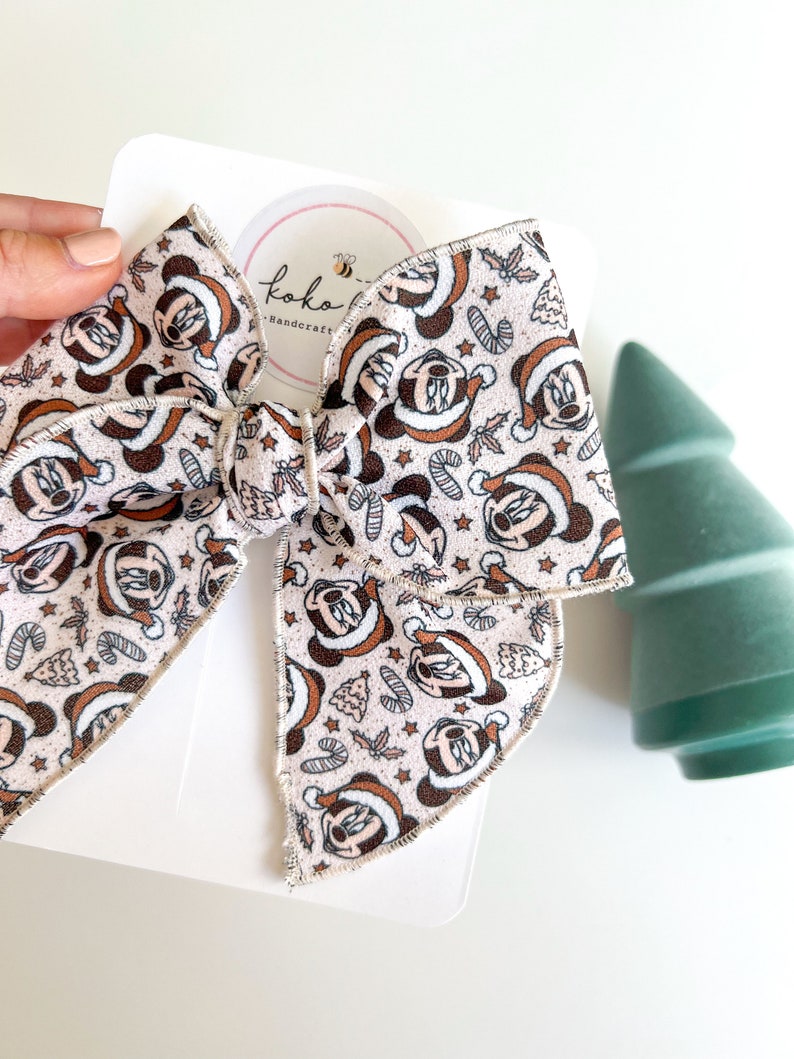 May include: A white fabric bow with a Christmas-themed Minnie Mouse print. The bow is tied with a white ribbon and has a stitched edge. The bow is on a white background with a small circle that says "Koko Handcraft" with a bee logo.