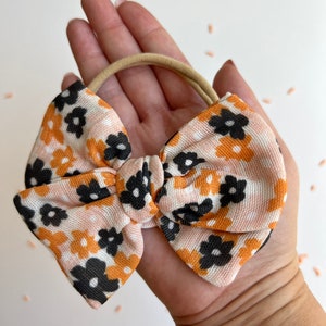 Op de afbeelding: Een haarstrik met perzik-, oranje- en zwarte bloemenpatroon. De strik wordt in een hand gehouden en heeft een beige elastiek. De strik heeft een herhalend patroon van kleine bloemen. De stof heeft een getextureerd uiterlijk.