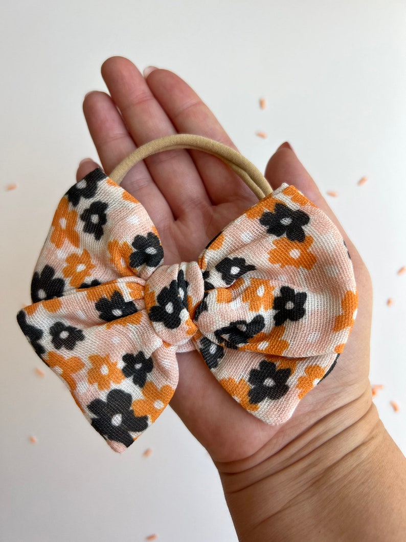 Op de afbeelding: Een haarstrik met perzik-, oranje- en zwarte bloemenpatroon. De strik wordt in een hand gehouden en heeft een beige elastiek. De strik heeft een herhalend patroon van kleine bloemen. De stof heeft een getextureerd uiterlijk.