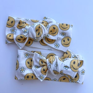 Pode incluir: Duas faixas de cabelo brancas com um padr&atilde;o repetido de rostos sorridentes amarelos e pequenas margaridas brancas. Cada faixa de cabelo tem um la&ccedil;o grande no centro. As faixas de cabelo s&atilde;o provavelmente para beb&ecirc;s ou crian&ccedil;as pequenas.