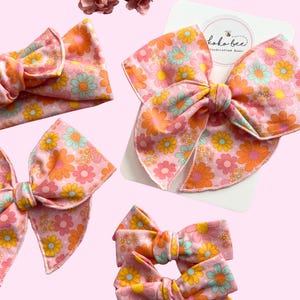 May include: A set of four pink and orange floral hair bows. The bows are made of fabric with a retro floral print. The bows are arranged in a variety of sizes and styles, including a headband, a large bow, and two smaller clips.