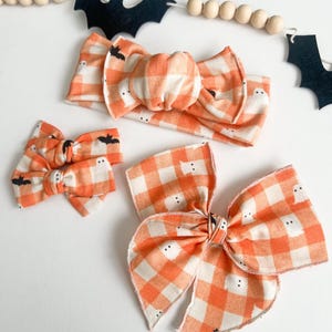 May include: Three Halloween-themed hair accessories: a headband and two bows. They feature an orange and white gingham pattern with small ghost and bat details. A wooden bead garland and felt bats are in the background.