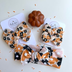 Op de afbeelding: Een set babyhaaraccessoires met een bloemenpatroon in zwart, bruin en oranje. De set bevat twee haarstrikken, een haarband en een kleine pompoendecoratie. De accessoires worden gepresenteerd op witte kaarten met de tekst "kokobee - handcrafted bows".