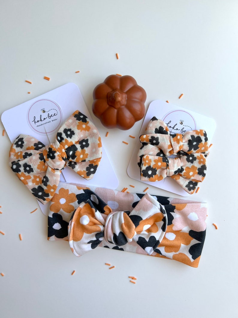 Op de afbeelding: Een set babyhaaraccessoires met een bloemenpatroon in zwart, bruin en oranje. De set bevat twee haarstrikken, een haarband en een kleine pompoendecoratie. De accessoires worden gepresenteerd op witte kaarten met de tekst "kokobee - handcrafted bows".