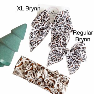 May include: Two Christmas-themed hair bows with a Mickey Mouse print in brown and white. The larger bow is labeled "XL Brynn" and the smaller bow is labeled "Regular Brynn". A matching headband with the same print is also shown.