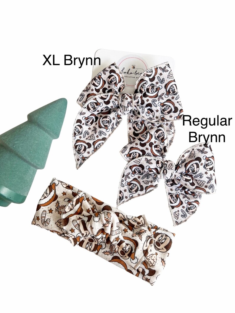 May include: Two Christmas-themed hair bows with a Mickey Mouse print in brown and white. The larger bow is labeled "XL Brynn" and the smaller bow is labeled "Regular Brynn". A matching headband with the same print is also shown.
