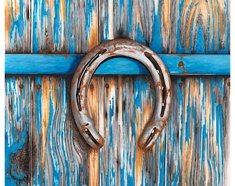May include: A metallic horseshoe with a weathered, rusted finish centered against a background of vertical wooden planks painted in distressed blue and natural tan tones. The text www.HenningArtStudio.com is visible at the top and bottom.
