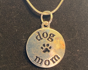 Puede incluir: Collar de plata con un colgante redondo que dice "dog mom" con una huella de pata negra en el centro.