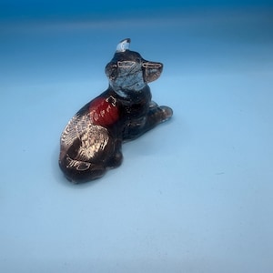 May include: A small ceramic figurine of a black and white dog with red accents. The dog is sitting with its head turned to the side.