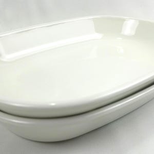 May include: Two white, oval-shaped ceramic serving dishes are stacked on top of each other. The dishes have a glossy finish and rounded edges. The image is taken on a white background.
