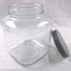 May include: A clear, square-shaped glass jar with a black lid. The jar has a wide opening and a textured design on the sides. The lid is off to the side.