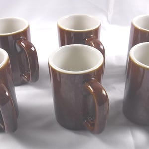 May include: Six brown ceramic mugs with white interiors and rounded handles are arranged on a white surface. The mugs have a glossy finish and a simple, classic design, suitable for coffee or tea.