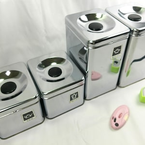 May include: A set of four chrome-finished canisters with black circular lids. The canisters are labeled "TEA" and "SUGAR" with small graphic icons. A pink mouse-shaped object and a green tape measure are also present.
