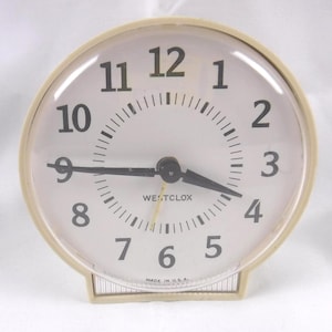 May include: A vintage, cream-colored Westclox alarm clock. The clock face is white with black numerals and hands. The clock reads approximately 8:40. The words "WESTCLOX" and "MADE IN U.S.A." are printed on the clock face.