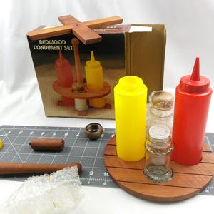 May include: A redwood condiment set with a wooden serving tray, two squeeze bottles, and two glass shakers. The set includes a yellow mustard bottle and a red ketchup bottle. The box reads "Redwood Condiment Set".
