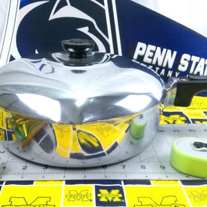 May include: A shiny, silver-colored cooking pot with a black handle and lid. The pot is on a patterned surface with a measuring tape nearby. A Penn State banner is in the background.