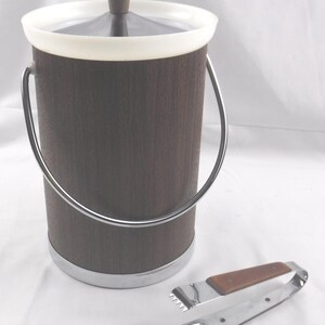 May include: A cylindrical ice bucket with a dark brown wood-grain exterior, a white rim, and a silver metal handle. The lid features a dark brown knob. A pair of silver metal ice tongs with brown wood-grain accents on the handles sits beside it.