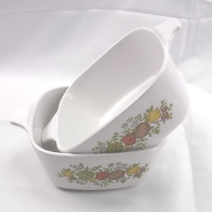 May include: Two white ceramic baking dishes with integrated side handles, stacked one inside the other. Each dish features a decorative botanical print along the exterior, displaying colorful illustrations of various vegetables and herbs.