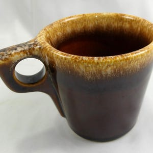 May include: A brown ceramic mug with a unique handle design. The mug features a gradient of colors, transitioning from a dark brown base to a lighter tan rim. The handle has a circular opening for easy gripping.