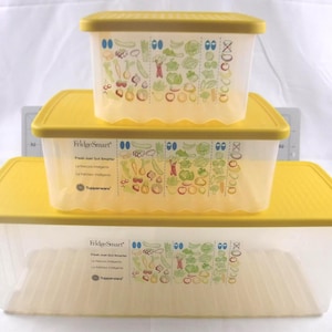 May include: Three clear plastic food storage containers with yellow lids are stacked. The containers have a rectangular shape and are labeled "FridgeSmart" with illustrations of vegetables. The containers are different sizes.