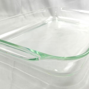 May include: A clear, rectangular glass baking dish with rounded corners and handles. The dish is empty and sits on a white surface. The glass has a slight green tint.