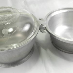 May include: Two vintage aluminum cooking pots. One pot has a clear glass lid with etched floral designs. The other pot is open, revealing a silver interior. Both pots have side handles for easy handling. The pots are on a white surface.
