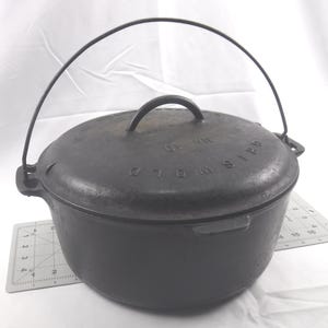 May include: A black cast iron Dutch oven with a lid and a wire handle. The lid has a small handle and the words "OLD WISHER" and "NO. 10" are embossed on the lid. The pot is sitting on a white surface.