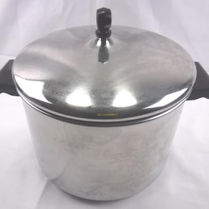 May include: A polished stainless steel stockpot with a matching lid. The pot features two black heat-resistant side handles, and the lid is topped with a black, faceted knob. The metal surface is reflective and smooth, showing a cylindrical shape.