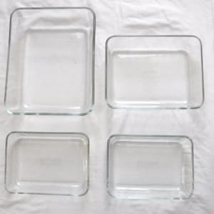 May include: Four clear glass rectangular baking dishes of varying sizes are arranged on a white surface. The dishes have rounded corners and are empty. The glass is transparent, allowing a view of the white background.