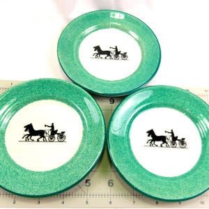 May include: Three decorative plates with a teal green rim and a white center. Each plate features a black silhouette of a horse-drawn carriage. The plates are arranged on a light-colored surface.