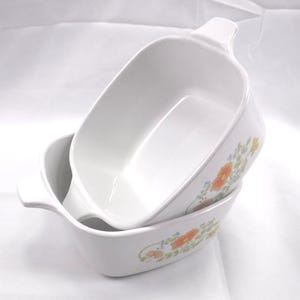 May include: Two white ceramic baking dishes with integrated side handles, stacked one inside the other. The exterior of the dishes features a floral pattern with orange flowers, green leaves, and small blue accents against a plain white background.