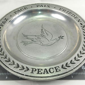 May include: A silver-colored plate with the word "PEACE" and other words in different languages around the rim. A dove carrying an olive branch is etched in the center, surrounded by a laurel wreath design.