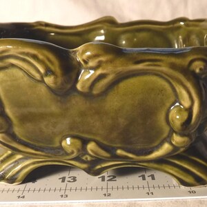 May include: A green ceramic planter with an ornate, raised design. The rectangular planter has a dark green interior and sits on four curved feet. The exterior features decorative scrollwork and a glossy finish, suitable for indoor plants.