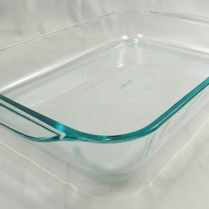 May include: A clear, rectangular Pyrex baking dish with rounded corners and teal-colored trim. The dish has a simple, functional design with a slightly raised rim and handles on either side. Ideal for baking casseroles, desserts, and more.