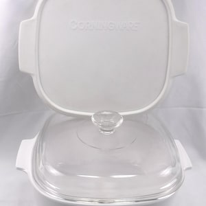 May include: A white square CorningWare casserole dish with integrated side handles and a clear glass lid featuring a central knob. A separate white square lid with the embossed text CORNINGWARE is positioned vertically behind the dish.