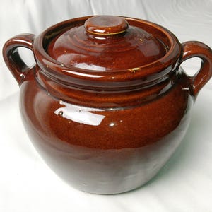 May include: A brown ceramic crock pot with a lid and two handles. The pot has a glossy finish and a rounded shape. The lid has a small knob on top.