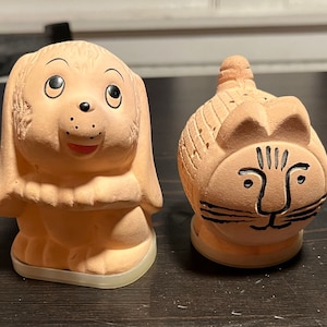 May include: A set of two ceramic salt and pepper shakers shaped like a dog and a cat. The dog is light brown with black eyes and a black nose. The cat is light brown with black stripes and a black nose.